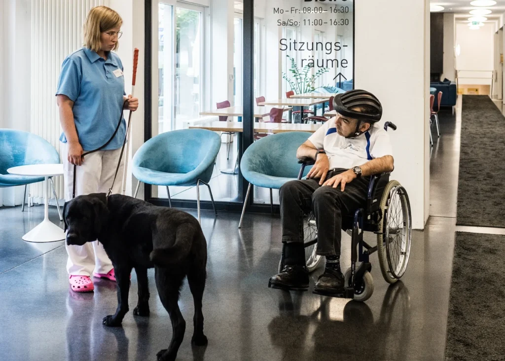 Mitarbeiterin mit Hund im Gespräch mit einem Bewohner, welcher im Rollstühl sitzt